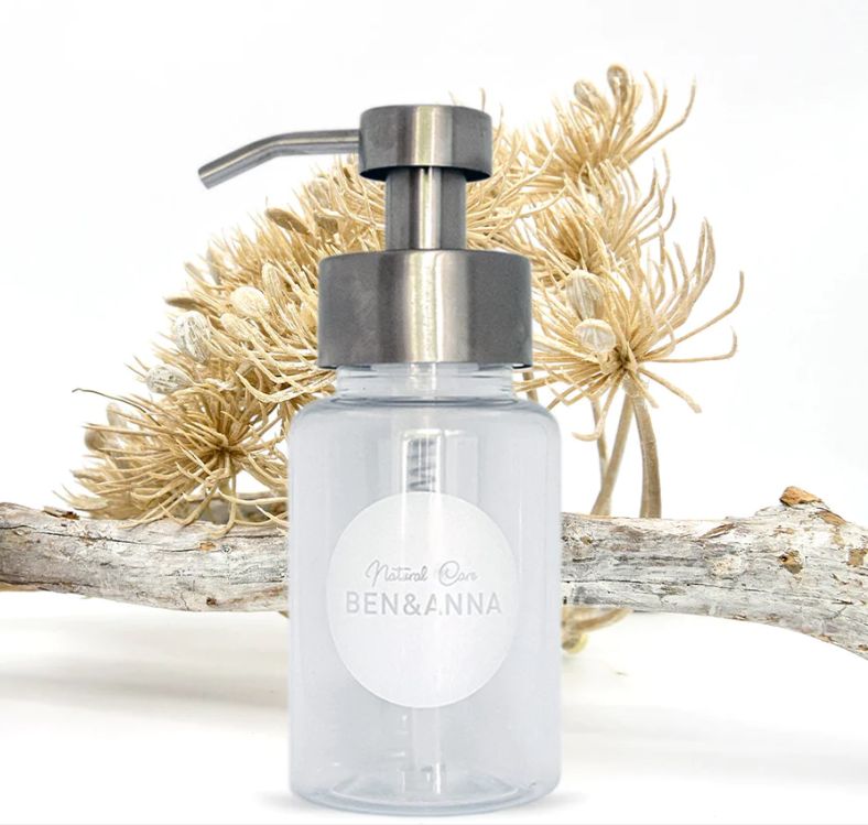 Transparante navulbare pompflacon van Ben & Anna met roestvrijstalen dispenser, gefotografeerd met natuurlijke decoratie op de achtergrond.
