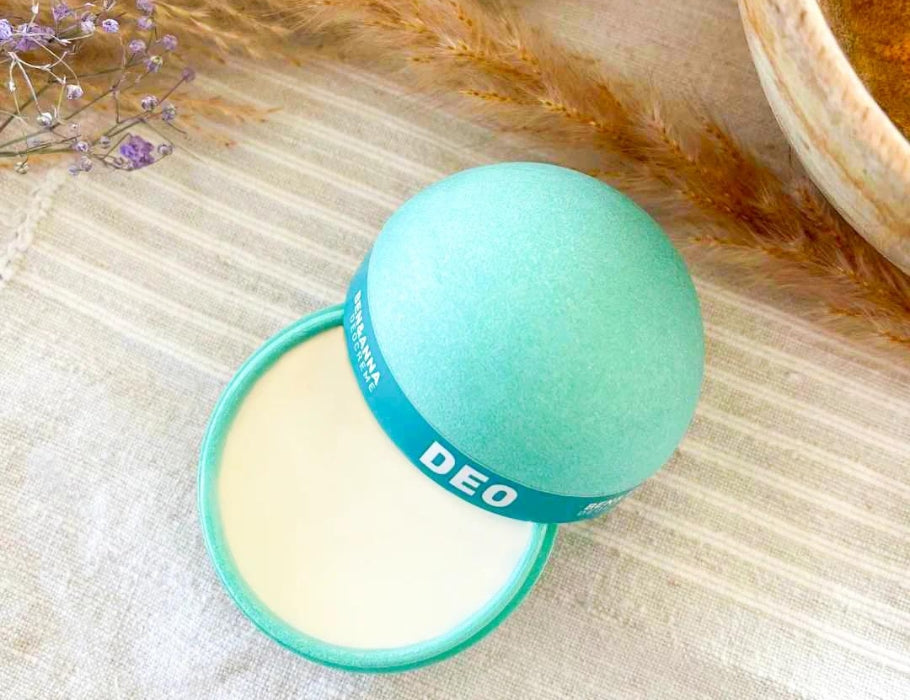 Open turquoise deodorantbal gemaakt van composteerbare PolyPotato®-verpakking, gevuld met een romige witte deodorantcrème. De balm ligt op een lichte stoffen ondergrond met gedroogde bloemen eromheen.