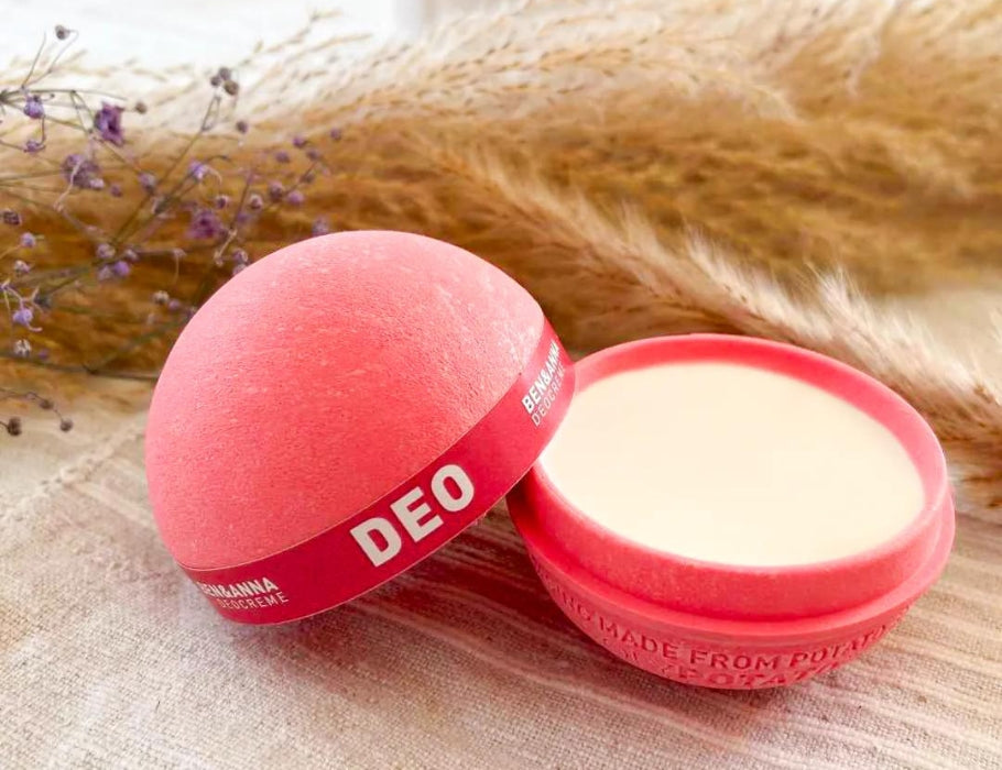Open roze Ben & Anna deodorantbal van composteerbare PolyPotato®-verpakking, met romige witte deodorant zichtbaar; liggend op een lichte stoffen ondergrond met gedroogde bloemen op de achtergrond.
