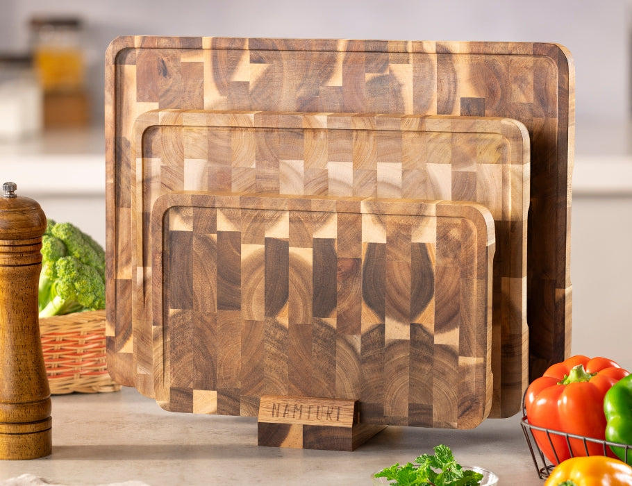 Set van drie Namture snijplanken van acaciahout in verschillende maten, rechtop geplaatst in een houten houder op een keukenblad, omringd door verse groenten.